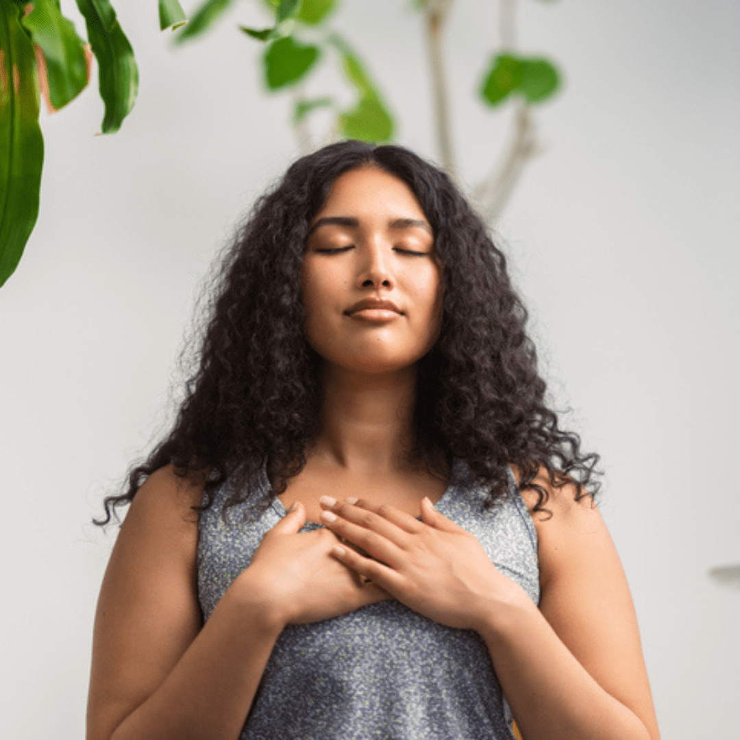 Multiracial woman in athleisure wear with her hands over her chest. The woman has a relaxed expression and is focused on breathing.